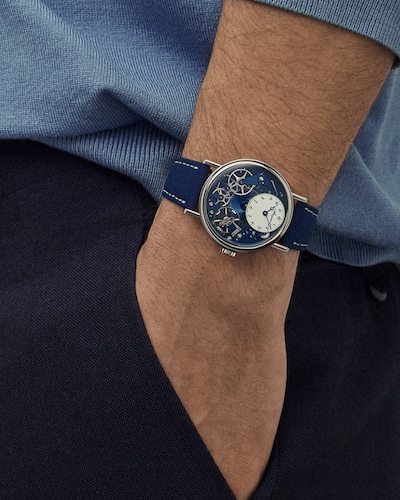 Two people wearing blue shirts and dark trousers stand against a neutral wall, each showcasing a stylish wristwatch with a blue strap and intricate dial design. image