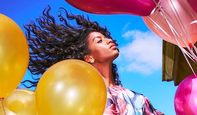 A person wearing Ted Baker earrings, surrounded by colorful balloons in shades of pink and gold, with vibrant curls blowing in the wind against a blue sky. image